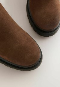 Punte di scarpe in suede marrone con suole in gomma nera su una superficie chiara, che mostrano dettagli di texture e materiale.