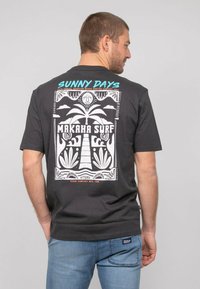 T-shirt en coton noir avec un design graphique blanc représentant des palmiers et un soleil, portant les inscriptions "JOURS EN SOLEIL" et "MAKAHA SURF". Manches courtes.