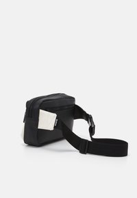 Borsa a vita bianca e nera con tasche con cerniera e tracolla nera regolabile, mostrata su uno sfondo bianco.