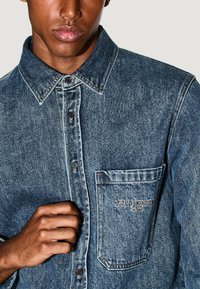 Camicia in denim blu con colletto a punta, taschino sul petto e branding ricamato. Presenta una chiusura standard e un tessuto testurizzato.