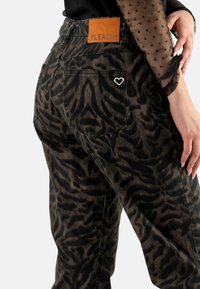 Pantalones con estampado de leopardo en marrón oscuro y negro, con un parche de cuero marrón, detalle de corazón plateado en la parte posterior y un corte relajado.