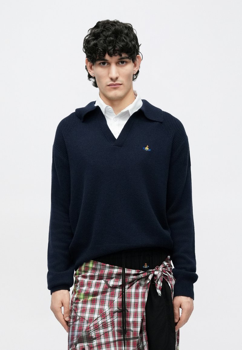 Jeune homme aux cheveux bouclés portant un pull col en V bleu marine par-dessus une chemise blanche et une jupe plissée à carreaux nouée sur un pantalon noir, debout devant un fond blanc.