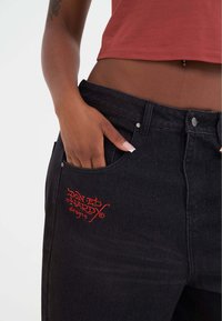 Černé džíny s přední kapsou, na které je výšivka červeného nápisu "Don Ed Hardy designs". Tkanina má hladkou strukturu a přiléhavý střih.