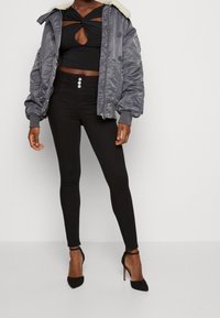 Grå bomberjacka med pälskrage, svart croptop med nyckelhålsskärning, högmidjade svarta leggings och svarta stilettohögder.