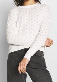 Weißer, gestrickter Pullover mit strukturiertem Muster, Rundhalsausschnitt und gerippten Bündchen und Saum. Über dunkelgrauen Jeans getragen.