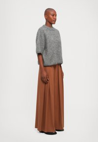 Femme avec la tête rasée portant un pull gris en peluche à manches courtes, un pantalon large plissé marron et des chaussures noires, se tenant de côté.