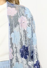 Průhledný kimono zdobený flitry s květinovými vzory v růžové, modré a tmavě modré barvě na lesklém stříbrném pozadí. Měkká textura.