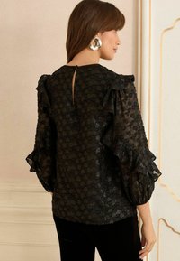 Svart blus med textur som har volangärmar, en nyckelhålbak och ett mönstrat design. Tyget har en genomskinlig kvalitet som förstärker den visuella djupet.
