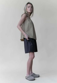 Beige mouwloze top, donkere shorts en grijze klompen. De top heeft een losse pasvorm, de shorts zijn knielang met een gestructureerde stof. Eenvoudig ontwerp.