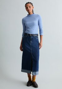 Femme debout portant un haut bleu à manches longues côtelées, une jupe midi en denim foncé avec une fente avant, une ceinture marron, des chaussettes moutarde et des mocassins marron foncé.