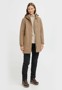 Braune, mittellange Jacke mit Kapuze, Reißverschluss und Seitentaschen. Kombiniert mit schwarzen Hosen und braunen Stiefeln mit roten Akzenten.