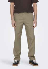 Beżowe spodnie o prostych nogawkach z trwałej tkaniny, wyposażone w rozporek na zamek, szlufki na pasek i gładką fakturę. Noszone z czarnymi trampkami.