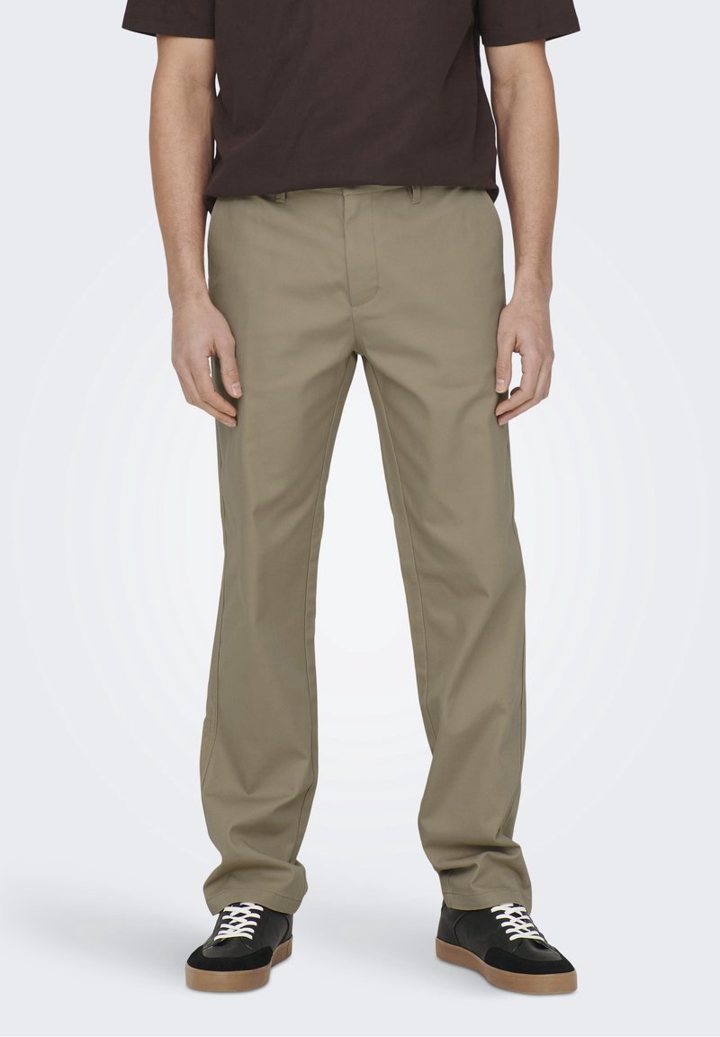 Beżowe spodnie o prostych nogawkach z trwałej tkaniny, wyposażone w rozporek na zamek, szlufki na pasek i gładką fakturę. Noszone z czarnymi trampkami.