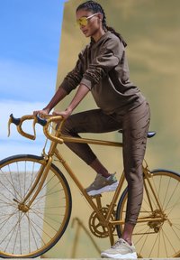 Brauner Hoodie mit strukturierten Details, passende braune Leggings mit verworrenen Mustern und beigefarbene Sneaker, die auf einem goldenen Fahrrad vor einem Hintergrund fahren.