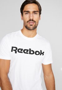 Hvit bomull T-skjorte med en fremtredende svart "Reebok"-logo over brystet, korte ermer og rund hals.