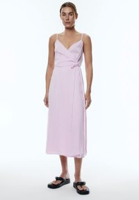 Helles rosa Wickelkleid mit dünnen Trägern, knielang, weiche Stofftextur, gebundenes Detail an der Taille, getragen mit schwarzen Sandalen.