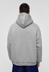 Grijze hoodie met een grote capuchon, ribgebreide manchetten en zoom. Zachte textuur, losse pasvorm, zichtbare stikdetails. Gekleed over donkere jeans.