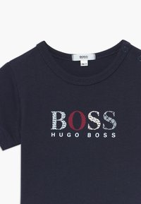 Námořnická modrá dětská tričko s krátkým rukávem vyrobené z měkké bavlny, které má multicolorové logo „BOSS“ a text „HUGO BOSS“ pod ním. Úchyty na rameni.