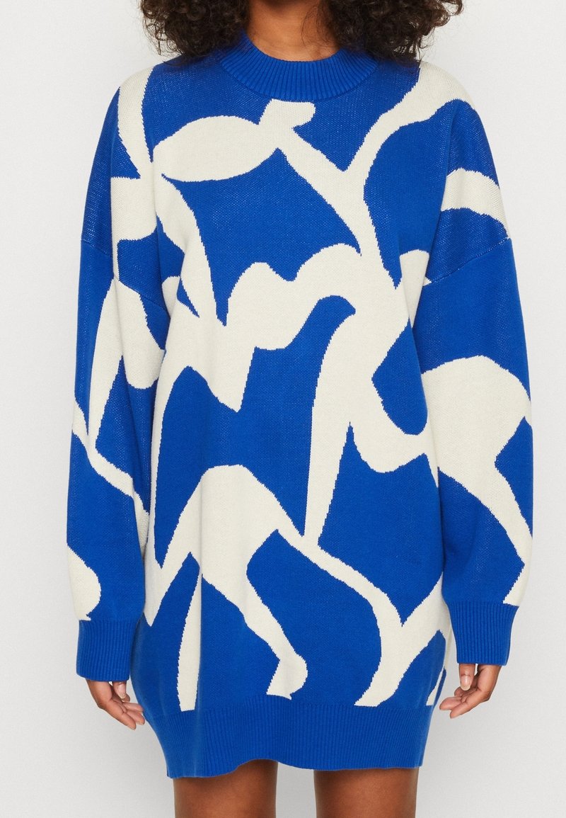 Robe-pull bleue surdimensionnée avec motif floral abstrait blanc, col rond côtelé, poignets et ourlet, portée par une personne aux cheveux bouclés.