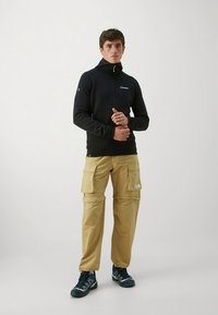 Μαύρο μπουφάν με φερμουάρ και κουκούλα, συνδυασμένο με μπεζ παντελόνι cargo με πολλές τσέπες και μπλε αθλητικά παπούτσια. Απλός, λειτουργικός σχεδιασμός.