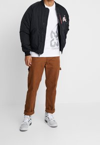 Chaqueta bomber negra con puños acanalados, camiseta gráfica blanca, pantalones cargo marrones y zapatillas grises con detalles en negro y blanco.