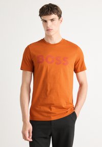 Oransje bomulls t-skjorte med rund hals, med ordet "BOSS" i en mørkere nyanse av oransje over brystet. Enkelt, korte ermer.