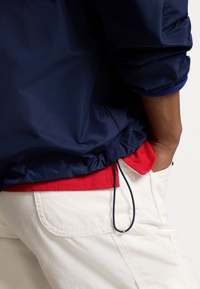 Coupe-vent marine avec des poignets élastiques et un ourlet froncé, superposé à une chemise rouge et un pantalon cargo blanc. Texture de tissu douce et lisse.