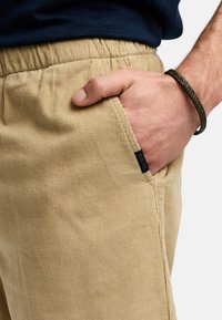 Beige kordfløjspants med en tekstureret overflade, elastisk talje og sidelomme. Et sort mærke er synligt nær lommen.