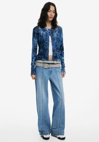 Cardigan à motifs bleus sur un top blanc, associé à un pantalon en denim bleu clair surdimensionné et une ceinture crème à double boucle. Chaussures noir et blanc.