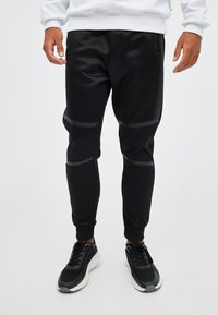 Pantalones deportivos negros hechos de tela suave, con detalles texturizados en los costados, puños ajustados y franjas horizontales a lo largo de los muslos.