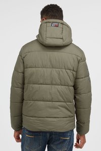 Uomo con giacca imbottita verde oliva con cappuccio e jeans blu, mostrato di spalle su uno sfondo semplice.