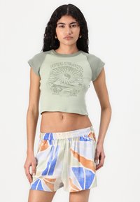 T-shirt cropped verde chiaro con stampa grafica, abbinato a pantaloni corti larghi e multicolore con motivi astratti arancioni, blu e verdi.