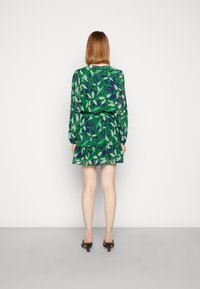 Lyhyt mekko, jossa vihreä lehtikuvio tummansinisen pohjalla, pitkät hihat, vyötäröltä kapeaksi puristettu ja kerroksittain laskostettu helma.