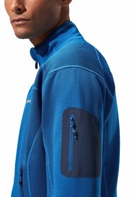 Blaue Sportjacke mit strukturiertem Muster, hohem Kragen und einer Reißverschlusstasche am Oberarm, die navyfarbene Akzente aufweist.