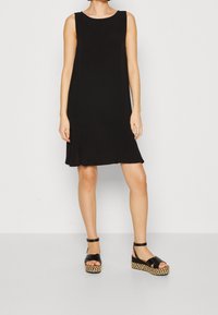 Robe noire sans manches avec une coupe décontractée et un ourlet évasé. Associée à des sandales à talons compensés noires avec une semelle tressée.