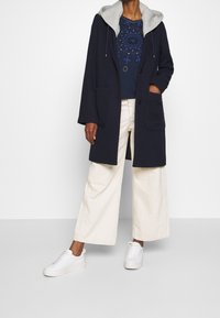 Marineblauer Mantel mit grauer Kapuze, zwei vorderen Taschen und Knopfverschluss. Getragen über einem marineblauen Hemd mit dekorativem Muster und cremefarbenen Hosen mit weitem Bein. Weiße Sneaker.