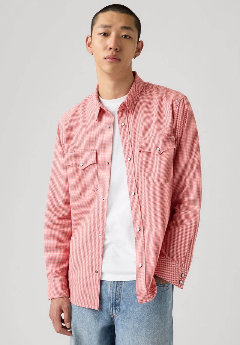 Giovane uomo che indossa una camicia a quadri rossa sopra una t-shirt bianca e jeans azzurri, in piedi contro uno sfondo semplice.