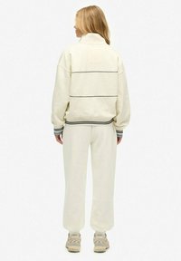Crème-kleurig trainingspak met een hoge kraag en gestreepte accenten op de manchetten. Losse pasvorm, zachte textuur, voorzien van een eenvoudig ontwerp en minimale details.