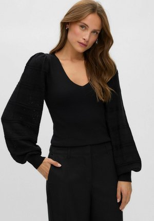 Femme aux longs cheveux bruns portant un haut noir à col en V avec de longues manches bouffantes et un pantalon noir, posant devant un fond uni.
