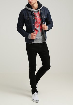Chaqueta de mezclilla en azul oscuro con herrajes plateados, superpuesta a una sudadera gris con impresión gráfica roja. Combinada con jeans negros de corte slim y zapatillas blancas.