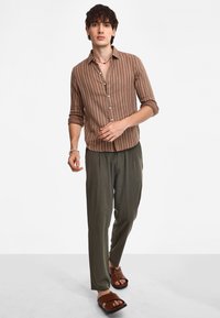 Camicia a righe marroni con bottoni, pantaloni leggeri verdi e sandali scivolosi marroni. La camicia ha le maniche lunghe arrotolate fino ai gomiti.