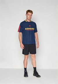 Ναυτικό αθλητικό μπλουζάκι με κόκκινες ρίγες και κίτρινο λογότυπο "ADIDAS", σε συνδυασμό με μαύρο σορτς. Το μοντέλο φορά μαύρα αθλητικά παπούτσια.