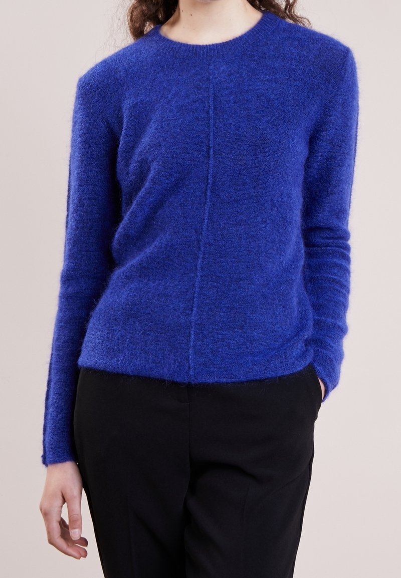 Personne portant un pull doux bleu roi à manches longues et un pantalon noir, une main dans la poche sur un fond uni.