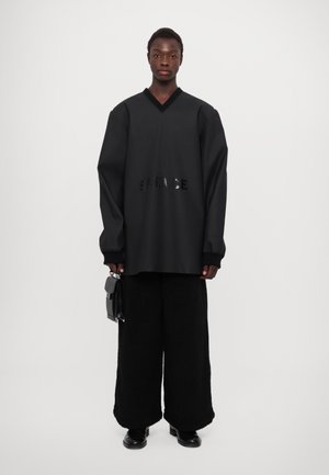 Hanorac oversized negru cu decolteu în V, prezentând un text „SILENCE” decupat. Asortat cu pantaloni lărgi negri texturați și pantofi negri lucioși.
