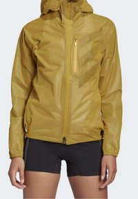 Lichtgewicht gele waterdichte jas met ritssluiting op zak, gestructureerde stof, verstelbare capuchon en elastische manchetten, gecombineerd met zwarte shorts.