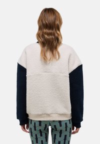 Veste en polaire avec un corps crème et des manches bleu marine, présentant une coupe décontractée et des poignets côtelés. Tissu texturé et confortable avec un design contrasté.