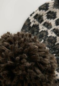 Knitted beanie in black, white, and brown with a textured leopard print design and a large, fluffy brown pom-pom on top.