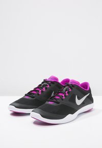 Scarpe sportive nere con accenti rosa e una suola bianca, dotate di un logo Nike argento, esposte su uno sfondo bianco uniforme.