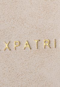 Lettere dorate in rilievo che formano la parola "XPATRI" su uno sfondo beige chiaro testurizzato, caratterizzato da un delicato motivo a sfumature.