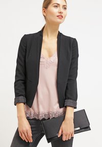 Fekete szabott blézer egy rózsaszín csipke camisole fölött, karcsú fekete farmerral és egy fekete táskával párosítva. Sima anyag, részletes csipke szegély.